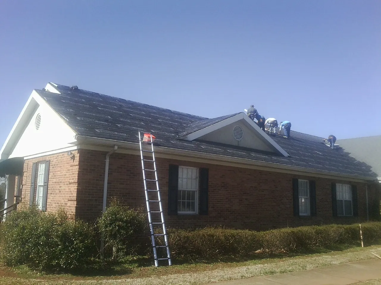 Crew tearing shingles off a brick ranch roof with a ladder set — Emergency Roof Repair work by our crew in Williamston